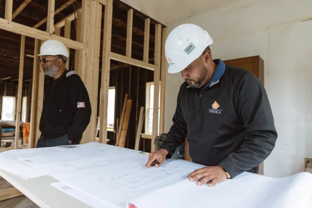 a person in a white hard hat looks at a big white paper on a table, while another person in a white hard hat stands to the side.